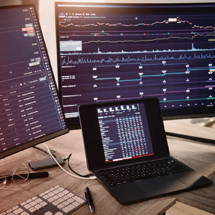 Multiple monitors and a laptop on a desk display real-time stock charts and data dashboards, representing the synergy between business, technology, and strategic financial analysis.