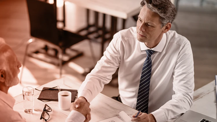 A professional handshake between a senior consultant and a client sitting at a desk, consolidating the start of a strategic collaboration and a relationship of trust.