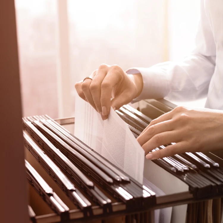 Close-up of hands browsing and selecting documents and hanging folders within an archive, illustrating the careful management and protection of governance frameworks and trust protocols.