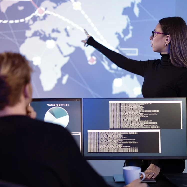 Analyst pointing to a global screen map while a colleague monitors computer vulnerabilities to strengthen fraud defense lines.